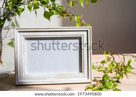 Portrait white picture frame mockup on wooden table. Modern glass vase with green branches. White wall background. Scandinavian interior. Eco lifestyle
