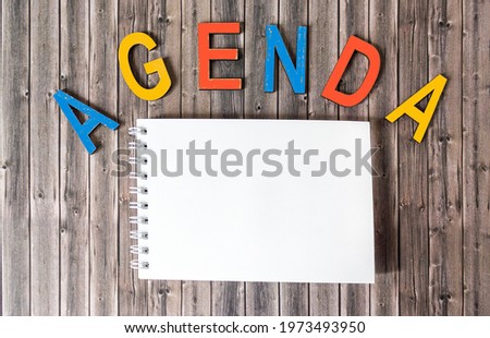 Of multicolored letters, the word AGENDA on a wooden table next to a blank notepad for writing