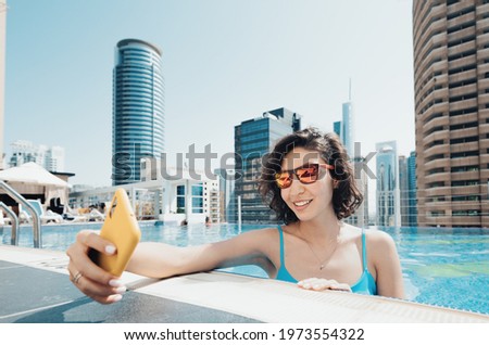 Cheerful woman travel blogger takes a selfie photo on her smartphone while relaxing in the pool on the roof of a skyscraper in Dubai