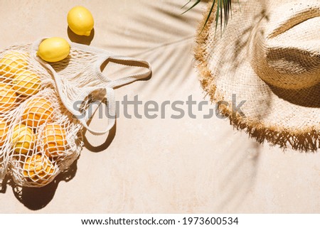 Summer flat lay on beige background. Straw hat and lemon fruits in eco friendly mesh shopping bag. Trendy palm shadow and sunlight, sun. Minimal summer travel fashion concept. Royalty-Free Stock Photo #1973600534