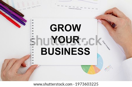 The girl with two hands holds a notebook with the text GROW YOUR BUSINESS, next to pencils, diagrams and graphs.
