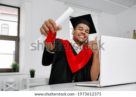 A laughing Black student in graduate gown and square cap who is happy to finish his studies shows his the long-awaited diploma, on Virtual graduation and convocation ceremony Class of 2021 Royalty-Free Stock Photo #1973612375