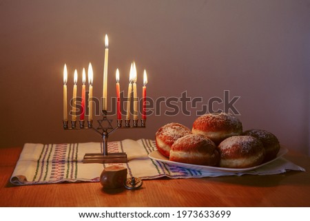 Menorah with lit burning candles for Jewish Hanukkah holiday on table at home. Celebrating Chanukah festival of lights. Dreidel and Sufganiyot donuts sweet cultural food on a plate. 