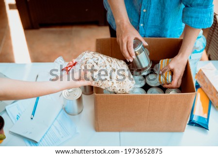 Volunteers collecting food into donation box. Working at food bank concept. Cut out middle section image of hands packing Cans at Food Drive. Royalty-Free Stock Photo #1973652875