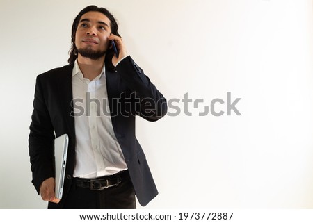 Hispanic man with long hair and a black suit holding a laptop and a smartphone on a white background