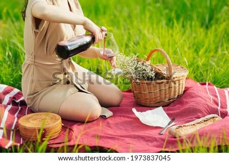 Cropped picture of a woman in dress pouring wine sitting on a green grass outdoors in the park. Concept of having picnic in a city park during summer holidays or weekends.