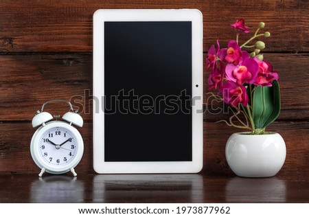 Tablet with Alarm clock and artificial flower decoration on a dark wooden shelf. Clipping path for tablet screen