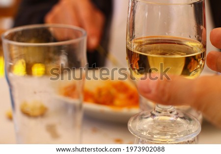 Close up of hand raising glass with white wine for a toast, photo taken at outdoor restaurant in front of a plate of pasta, focus on the glass, background out of focus.