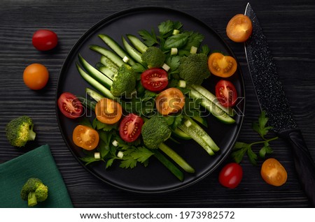 A black round plate of fresh vegetables stands on a black wooden table with a black knife, tomatoes and broccoli nearby. Top view. 