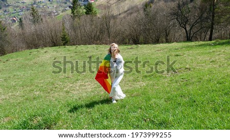 Young blonde woman running with LGBT rainbow flag in the mountains on nature in white wedding dress. Happy girl celebrates a special day