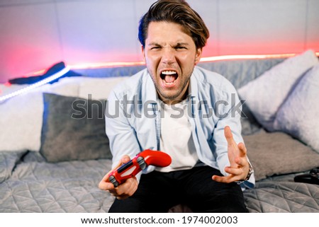 Photo of handsome excited guy sitting at comfy sofa, quarantine stay home gamer holding play station joystick, good mood fifa championship, Unhappy gamer player dissatisfied with video game. Royalty-Free Stock Photo #1974002003