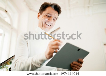 A confident composer does the work in a tablet. A gray-haired, positive man in age, writes down new cases on the project in the gadget. A close portrait of a creative person with white teeth.