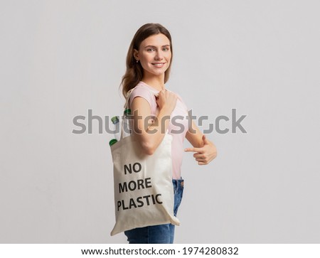 Beautiful Millennial Female Carrying Eco Tote Bag With No More Plastic Inscription Filled With Empty Bottles And Pointing On It, Volunteer Lady Posing Over Light Studio Background, Copy Space Royalty-Free Stock Photo #1974280832