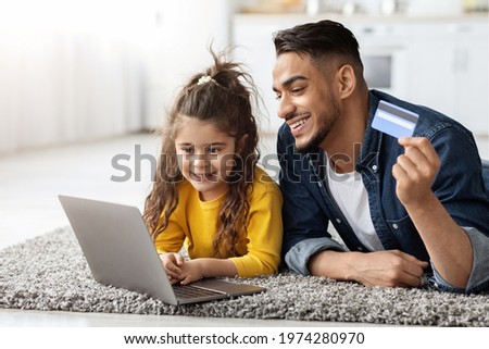 Portrait of happy arab dad and cute little daughter with laptop and credit card making online shopping together at home, middle eastern family relaxing on carpet with computer, closeup shot