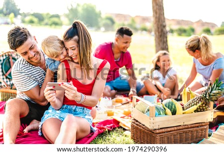 Happy multiethnic families playing with phone at pic nic garden party - Joy and love life style concept with mixed race people having fun together at picnic barbecue before sunset - Warm bright filter