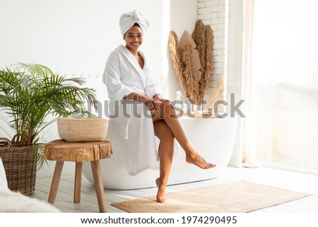 Wellness And Spa. Attractive Black Lady In White Dressing Gown Sitting On Bathtub Touching Smooth Legs After Depilation In Bathroom. Female Caring For Body And Skin At Home. Bodycare And Skincare Royalty-Free Stock Photo #1974290495