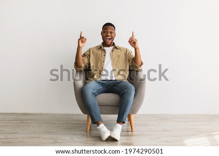 Excited African American Millennial Man Pointing Fingers Up Sitting In Chair Over Gray Wall Background, Looking At Camera. Check This Our, I Have Great Idea Concept. Front View Royalty-Free Stock Photo #1974290501