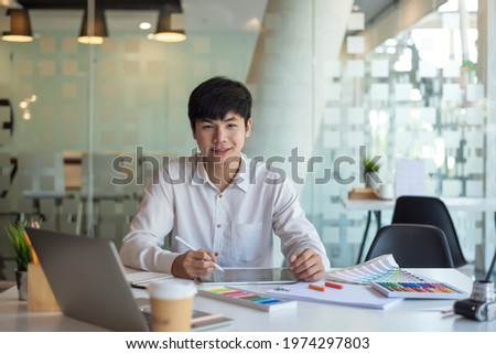 Young Asian designer man handsome sitting at work looking at the camera with his work equipment placed at a desk in the office.