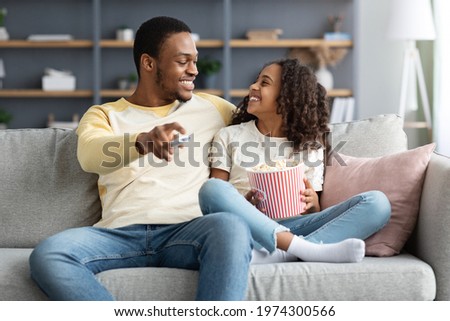 Adorable black family father and daughter watching TV together at home. Smiling african american dad and school girl watching funny cartoon and eating popcorn, sitting on couch, copy space