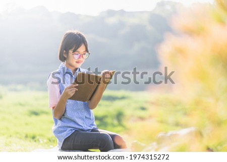 Cute asian little girl with glasses reading a book in garden at summer time