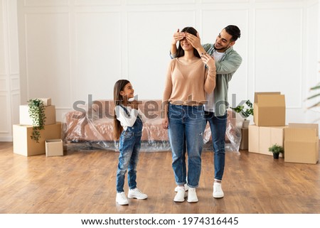 Moving New Home. Full Body Length Of Happy Man Covering His Wife's Eyes Before Showing Their Apartment After Relocation Standing Indoor With Their Excited Little Daughter. Free Space Royalty-Free Stock Photo #1974316445