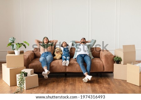 Moving Day. Lovely arab family relaxing on the couch in new home with cardboard boxes around. Happy man, woman in headscarf and little girl lying on sofa and looking up, free copy space Royalty-Free Stock Photo #1974316493