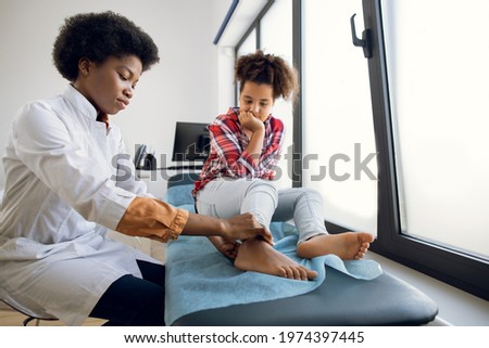 Cute curly mixed race teen 12-aged girl, sitting on the couch at modern clinic, while young 30-aged female African doctor orthopedist rheumatologist examining patient's leg Royalty-Free Stock Photo #1974397445