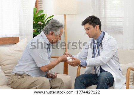 Doctor hold bottle of pills and explaining using medication to Asian elderly man patient sitting on sofa in living room after medical examination. Doctor provided the service to visit at home.