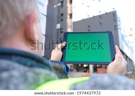 A man holds a tablet with a green screen in his hands against the backdrop of a building under construction. Architect or designer