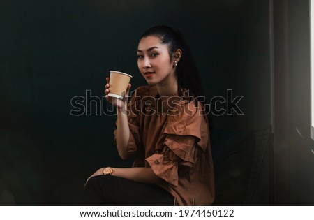 Enjoying an asian woman drinking hot coffee from a paper cup, she sits comfortably next to a window with soft light outside : The lifestyles of cute Thai girls love drinking coffee.