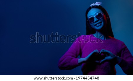 Color light portrait. Love sign. Romantic sympathy. Blue neon supportive grateful smiling Asian fan girl in pink showing heart gesture isolated on dark night copy space background.