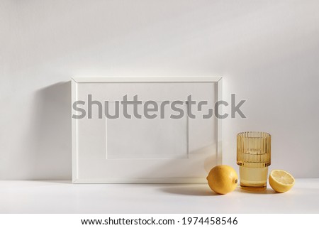 Summer stationery still life scene. Cut lemon fruit. Golden rippled glass of water on beige table background in sunlight. Blank white horizontal picture frame mockup. Champagne wall, long shadows.