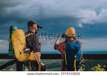 Couple of young hikers taking photo with a smartphone and looking into nature with a binoculars while standing on a lookout