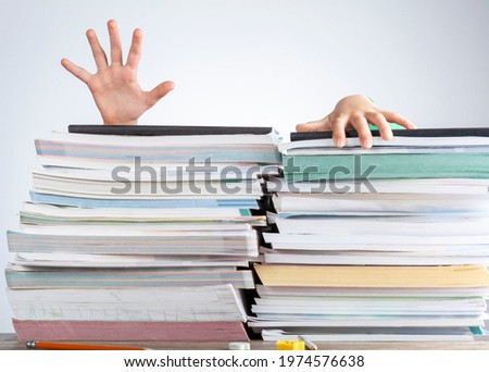 Abstract concept image showing a young student behind a large pile of test prep books on a study desk. An overwhelming load. The kid is trying to escape by climbing onto the pile as if she is drowning Royalty-Free Stock Photo #1974576638