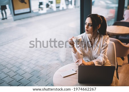 Young serious woman in white blouse with hair tied up looking out picture window and drinking coffee while working in sidewalk cafe