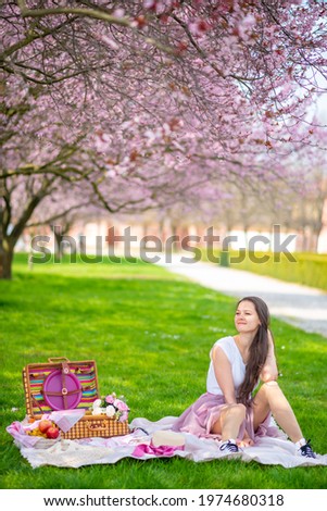 Beautiful woman in straw hat under blooming cherry trees in picnik place. Girl wearing a pink long skirt in spring garden