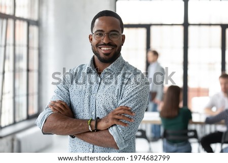Portrait of happy African American small business owner posing with hands folded. Millennial black male team leader smiling, looking at camera, employees working in modern office behind. Head shot Royalty-Free Stock Photo #1974682799