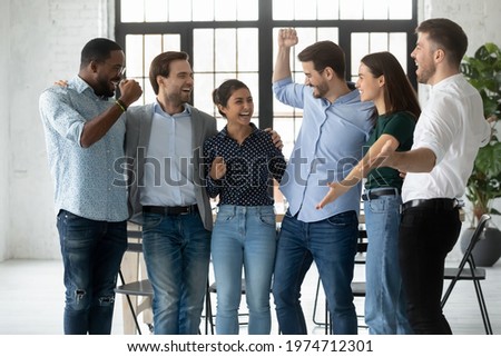 Excited motivated colleagues shouting for joy and celebrating team success in office. Happy employees hugging, dancing, making win gestures, laughing and having fun together. Unity, teamwork concept Royalty-Free Stock Photo #1974712301