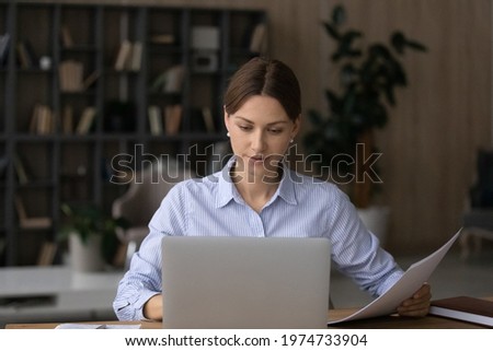 Computer and papers. Focused millennial woman office worker teacher freelancer do paperwork manage legal documents fill in electronic form. Young lady lawyer study text of contract agreement by laptop Royalty-Free Stock Photo #1974733904