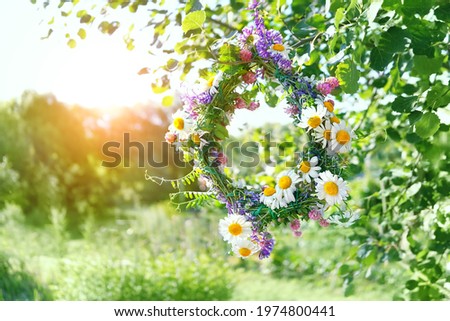 beautiful wreath of meadow flowers hanging on tree, natural sunny green background. floral decor, symbol of Summer Solstice Day, Midsummer. pagan witch traditions, wiccan ritual. Litha sabbat. Royalty-Free Stock Photo #1974800441
