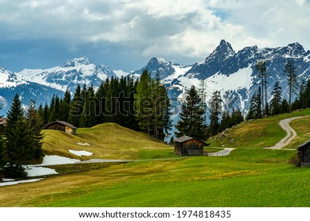 little alp with flowered meadows and black forests under the incoming Storm front, over the snowy mountains lies thunderstorm mood, sun rays break through the clouds and illuminated alps, foehn scene Royalty-Free Stock Photo #1974818435