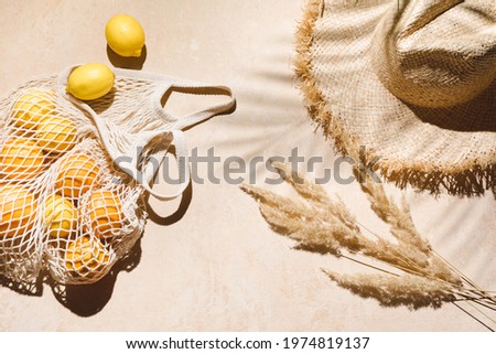 Summer flat lay on beige background. Straw hat and lemon fruits in eco friendly mesh shopping bag. Trendy palm shadow and sunlight, sun. Minimal summer travel fashion concept. Royalty-Free Stock Photo #1974819137