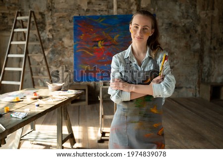 Satisfied female artist posing near new picture next to easel. Portrait of a female artist with crossed arms
