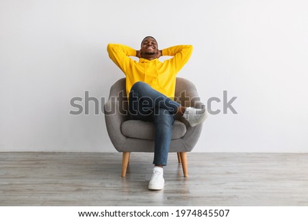 Lazy Weekend. African American Guy Relaxing With Eyes Closed Holding Hands Behind Head Sitting In Armchair Over Gray Wall Background, Wearing Yellow Hoodie. Relaxation And Comfort Concept Royalty-Free Stock Photo #1974845507