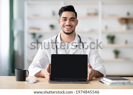 Cheerful middle-eastern man doctor showing laptop with blank screen while sitting at workplace in hospital or at home, copy space, mockup. Remote healthcare, distance medicine concept