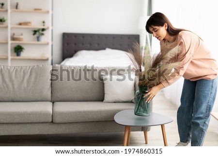 Modern Decoration Concept. Casual young lady putting glass vase with dried flowers on tea table. Millennial woman decorating her modern apartment and bedroom or living room with pampas grass Royalty-Free Stock Photo #1974860315