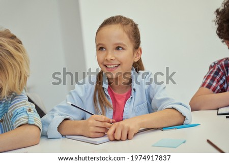 Lovely little schoolgirl smiling away, holding pencil and listening while studying, sitting at the table in elementary school classroom