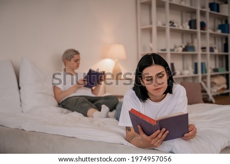 tattooed woman lying on bed and reading book near girlfriend on blurred background