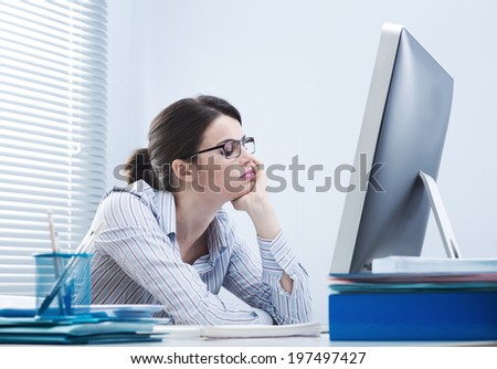 Bored office worker at desk staring at computer screen with hand on chin.