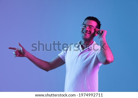 Pointing at side. Latina young man's portrait on purple-blue studio background in neon light. Handsome male model in casual style. Concept of human emotions, facial expression, youth, sales, ad.